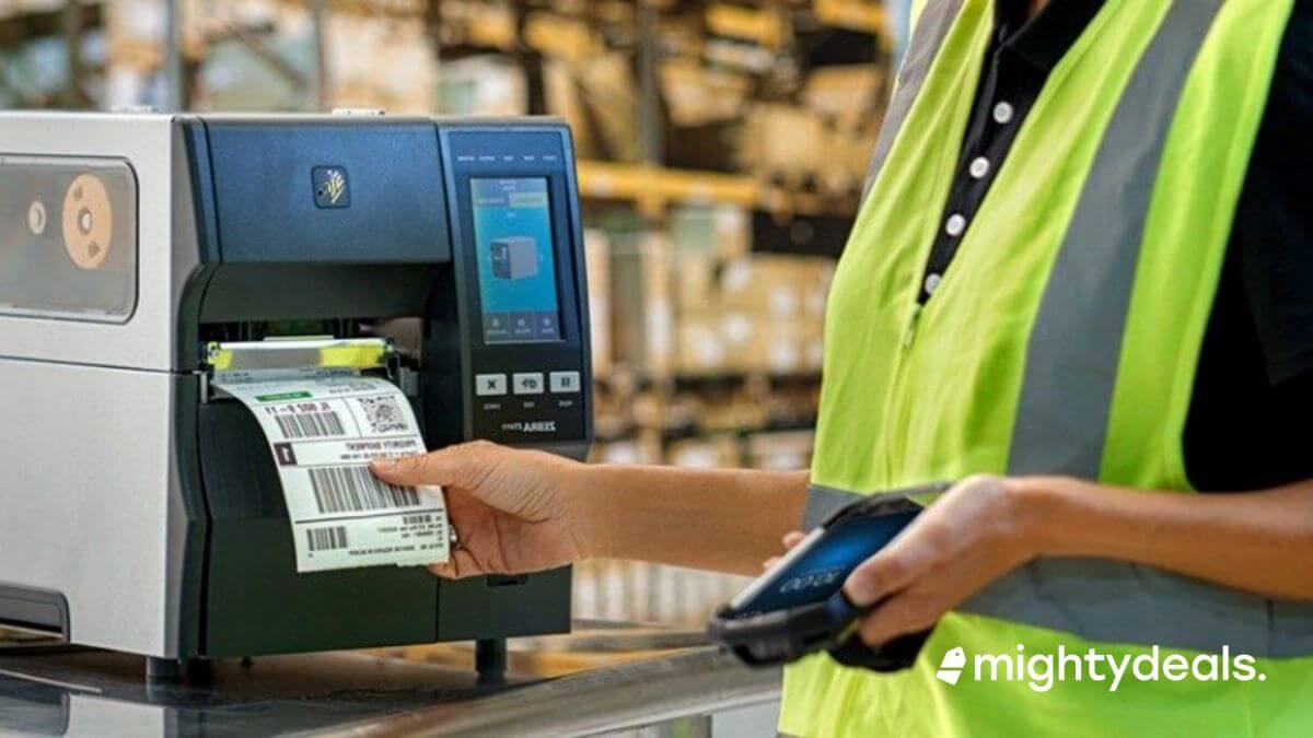 An industrial label printer is situated on the right, featuring a touchscreen interface and a blue and black color scheme. A label with a barcode is being printed and collected by a person on the left. The person is partially visible, wearing a safety vest with fluorescent green and a black undershirt. They are also holding a scanning device in their left hand, which is resting on a dark surface, possibly a conveyor or a workbench. The background is softly focused, hinting at an industrial setting with warehouse shelves. The "mightydeals" watermark suggests a promotional or commercial context for the image.